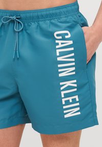 Aqua spalvos maudymosi šortai, pagaminti iš lygaus audinio, su raišteliu juosmenyje ir vertikaliu baltu "CALVIN KLEIN" logotipu šone.