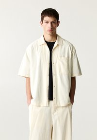 Camicia a maniche corte di un leggero beige, con bottoni e tessuto texturizzato, con una tasca frontale, indossata sopra una maglietta nera. Design del colletto classico.