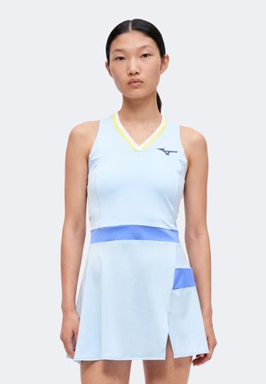 Jeune femme portant une robe de tennis sans manches bleu clair avec un col rayé jaune et des accents bleus à la taille et sur les poches, debout devant un fond blanc.
