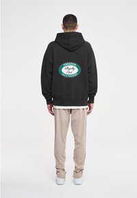 Hombre de espaldas con sudadera negra con el logo "Pegador Royalty Club Cold Hearted", pantalones beige y zapatillas blancas sobre fondo blanco.