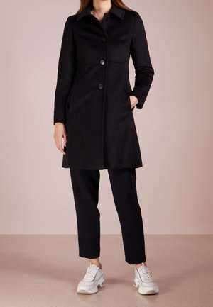 Kvinde iført en sort knapjakke, sorte bukser og hvide sneakers, stående med den ene hånd i jakkelommen mod en ensfarvet baggrund.