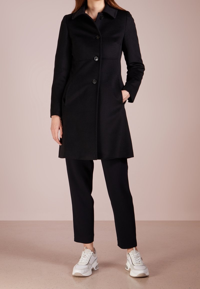 Femme portant un manteau noir boutonné, un pantalon noir et des baskets blanches, debout avec une main dans la poche du manteau devant un fond uni.