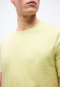 Νεαρός άνδρας που φοράει ανοιχτό κίτρινο T-shirt με στρογγυλή λαιμόκοψη και διακριτικό ανάγλυφο λογότυπο BOSS στο στήθος, κοιτάζοντας ελαφρώς προς τα αριστερά.