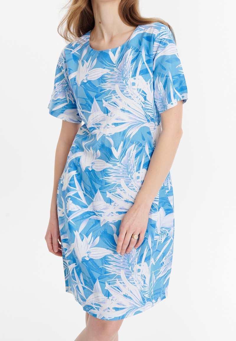 Femme portant une robe à manches courtes, longueur genou, avec des motifs de feuilles tropicales bleues et blanches, debout devant un fond blanc uni.