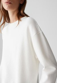 Donna che indossa un maglione a maniche lunghe in maglia waffle bianco con scollo rotondo, mostrata dalla metà del busto al mento su uno sfondo semplice.