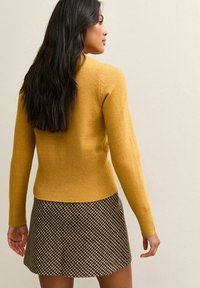 Geel wollen trui met lange mouwen en een aansluitende taille, gecombineerd met een korte, geruite rok in bruin en beige kleuren.