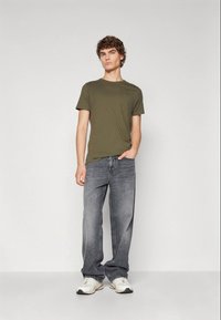 Olive green cotton t-shirt paired with loose-fitting gray jeans and white sneakers, standing against a neutral background.