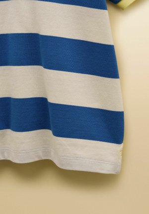 Sinise ja valge horisontaalselt triibulise kootud kangas nähtava tekstuuriga, veidi kaarekujuliselt riputatud beeži tausta ees.