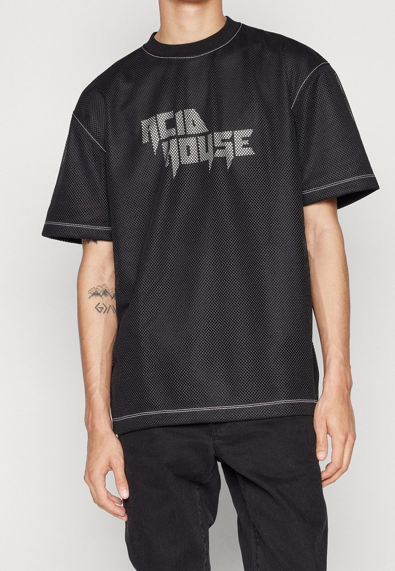 Svart mesh T-shirt med korta ärmar, med ett grafiskt tryck "ACID HOUSE" i silver och kontrasterande vit sömnad på kanterna.