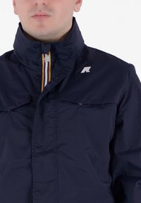 Giacca blu navy con colletto alto, zip argentata, due tasche sul petto e un piccolo logo sul lato sinistro. Tessuto liscio e leggero.