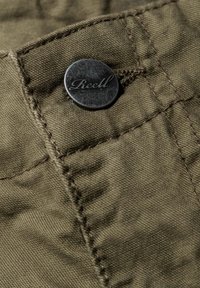 Nahaufnahme von olivgrünem Stoff, der einen Metallknopf mit eingraviertem "Reell", strukturiertem Nähen und einer glatten Oberfläche zeigt.