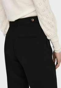 Pantalones negros ajustados con una textura suave, con cierre de botón y dos bolsillos traseros. La cinturilla se sitúa justo por encima de las caderas.
