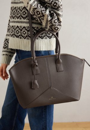 Personne portant un pull à motifs et un jean tenant un grand sac à main en cuir marron foncé avec double poignée et fermeture éclair.