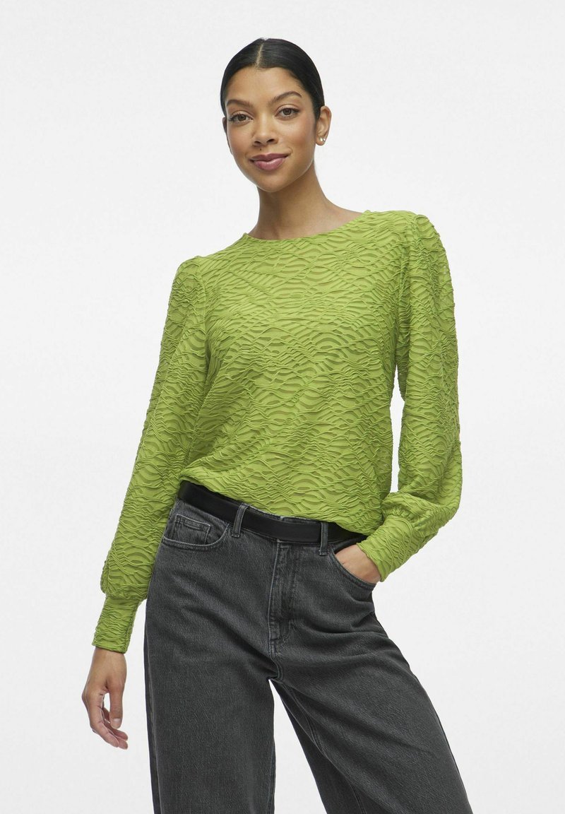 Haut à manches longues texturé de couleur vert vif avec un motif en vagues, associé à un jean foncé taille haute et une ceinture noire.