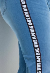Jeans de mezclilla azul con una franja lateral que lleva el texto "BIG DREAM" en negro y blanco, resaltado por un contorno rosa.