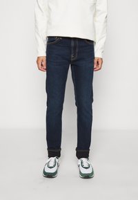 Calças de ganga azul-escura com um corte slim e design tradicional de cinco bolsos, apresentando costuras amarelas contrastantes e detalhes sutis de desgaste.