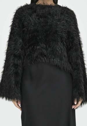 Femme portant un pull court noir à manches longues avec une texture duveteuse, sur une jupe noire unie, debout devant un fond neutre.