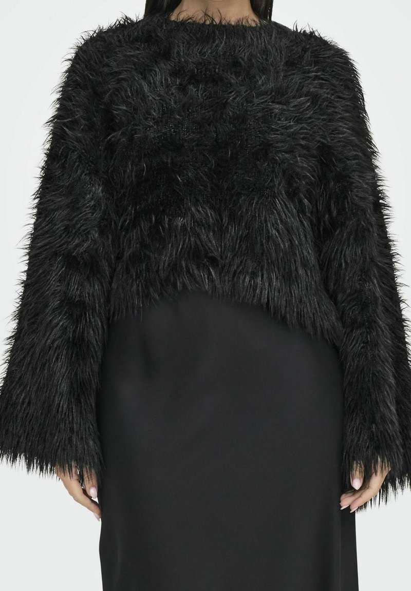 Femme portant un pull court noir à manches longues avec une texture duveteuse, sur une jupe noire unie, debout devant un fond neutre.