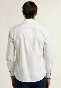 Camisa blanca de botones con mangas largas, textura suave, dobladillo curvado y acentos azules en contraste en los puños. Vista trasera.