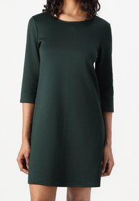 Nainen, jolla on yllään tummanvihreä, polvipituinen mekko, jossa on kolmanneksen pituiset hihat ja pyöreä kaulus, seisoo valkoista taustaa vasten.