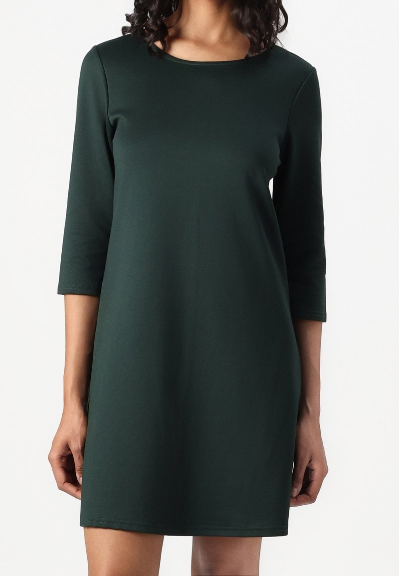 Nainen, jolla on yllään tummanvihreä, polvipituinen mekko, jossa on kolmanneksen pituiset hihat ja pyöreä kaulus, seisoo valkoista taustaa vasten.