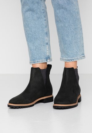 Svarta mocka ankelskor med elastiska sidopaneler, en gummisula och en beige kant längs kanten. Bärs med ljusblå jeans med uppvikte ben.