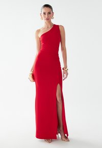 Červené večerné šaty na jedno rameno s vysokým rozparkom. Vyrobené z hladkého materiálu s tesným strihom. Doplněné zlatými šperkami a kabelkou.