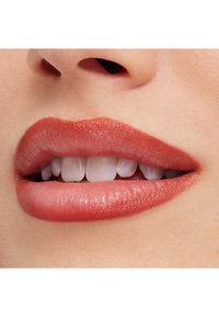 Zbliżenie na wargi pokryte brzoskwiniowo-różową matową szminką. Gładka tekstura, delikatny połysk na górnej wardze, białe zęby widoczne między rozchylonymi wargami.