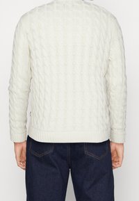 cremefarbener, kabelgestrickter Pullover mit langen Ärmeln, geripptem Halsausschnitt und Bündchen sowie strukturierter Oberfläche. Getragen mit dunkelblauen Jeans.