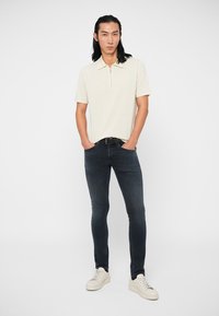 Ανοιχτό μπεζ πόλο μπλουζάκι με φερμουάρ στον λαιμό, συνδυασμένο με σκούρα μπλε στενά τζιν και λευκά αθλητικά παπούτσια. Απλός, καθαρός σχεδιασμός με ελάχιστες λεπτομέρειες.