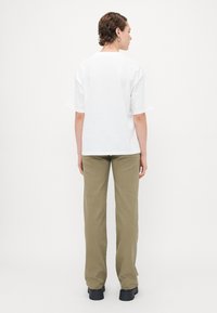 Person med kort lockigt hår som bär en enkel vit oversized t-shirt, olivgröna raka byxor och svarta klumpiga skor, sedd bakifrån.