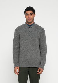 Γκρι πλεκτή μπλούζα με γιακά, τρία κουμπιά και υφή ύφανσης. Μακριά μανίκια και άνετη γραμμή, φορεμένη πάνω από μπλε πουκάμισο.