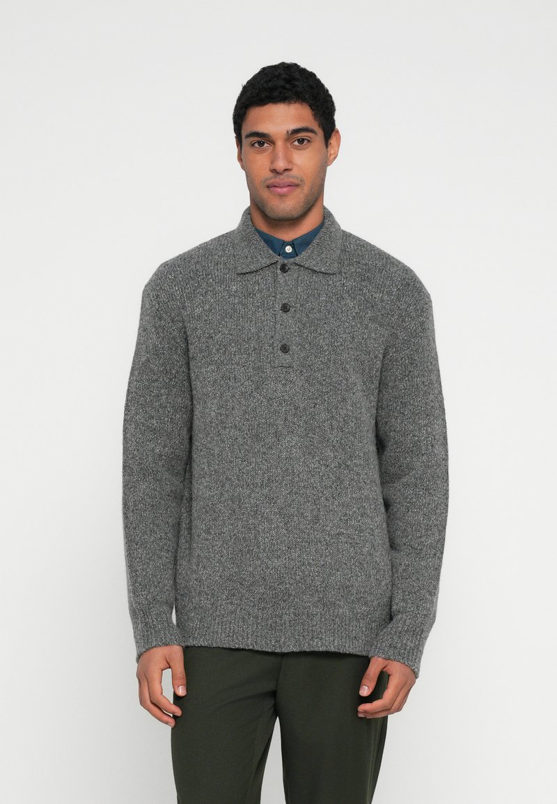 Γκρι πλεκτή μπλούζα με γιακά, τρία κουμπιά και υφή ύφανσης. Μακριά μανίκια και άνετη γραμμή, φορεμένη πάνω από μπλε πουκάμισο.