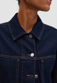 Giacca di jeans blu scuro con colletto, bottoni in argento e cuciture arancioni a contrasto. Primo piano della consistenza del tessuto e dei dettagli.