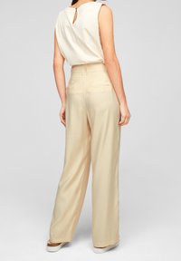 Beiga byxor med vida ben och en mjuk textur, med en dragkedja bak och två bakfickor. Tyget faller mjukt.