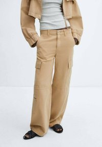 Beige cargo bukser med bred bendesign, flere sidelommer og en subtil tekstur. Parret med en tætsiddende grå top og sorte sandaler.