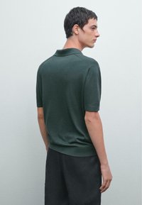 Tmavě zelené polo tričko s krátkými rukávy, žebrovaným límečkem a lemem. Hladký povrch a přiléhavý střih. Model stojí zády, má na sobě černé šortky.
