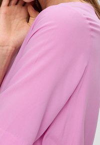 Tissu léger rose avec une texture lisse. Le vêtement présente une coupe décontractée et un col arrondi. Gros plan sur le détail de la manche visible.