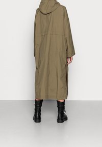 Abrigo largo color verde aceituna con capucha, hecho de material ligero. Presenta aberturas laterales y un corte holgado. Combinado con botas de combate negras.