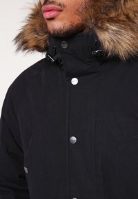 Černá parka s kapucí lemovanou umělou kožešinou, se sklapovacími knoflíky, hladkým povrchem a předními kapsami.