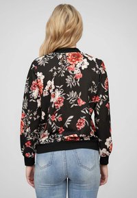 Veste bomber florale avec un fond noir, ornée de fleurs rouges et blanches, avec des poignets et un ourlet côtelés, ainsi qu'une texture lisse et brillante.