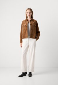 Femme debout dans une veste marron à boutons, un haut blanc, un pantalon large crème, des bottes noires et des lunettes, sur un fond blanc uni.