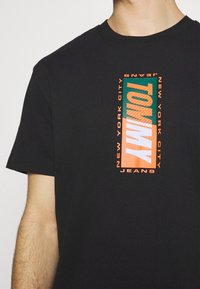 Mustat puuvillaiset t-paidat, joissa on suorakulmainen monivärinen graafinen kuva, jossa on teksti "TOMMY", "NEW YORK CITY" ja "JEANS" -korostuksia. Lyhyet hihansuut.