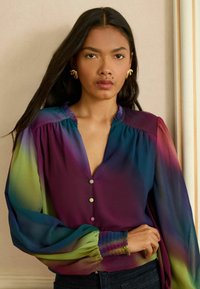Bluzka o luźnym kroju, z gradientem fioletowego, zielonego i niebieskiego, z dekoltem w kształcie litery V, detalami guzików oraz ściągaczami przy mankietach.