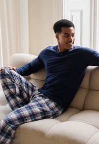 Marineblauwe lange mouwen shirt met een knoopdetail, gecombineerd met blauw-witte geruite pyjamabroek op een beige bank. Zachte stof, relaxte pasvorm.