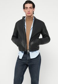 Fekete bőrból készült bomberdzseki elöl cipzárral, oldalzsebekkel és testhezálló kialakítással, barna garbó és világoskék ing rétege fölött viselve.