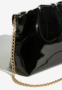 Melna lakādas rokassoma ar viļņotu dizainu, zelta ķēdes siksniņu un leņķa formu pamatni. Gluda, spīdīga tekstūra un izteiksmīga silueta.