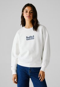 Sudadera blanca con el logo de "Red Bull Racing" en color marino. Presenta un cuello redondo y puños acanalados. Combinada con jeans de mezclilla azul.