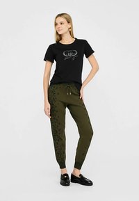 Must t-särk sädelevate logodega, kombineeritud oliivroheliste jopedega, millel on tume leopardimuster. Modell kannab musti slip-on kingi.