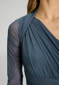 Sininen, hihaton mekko, jossa on laskostettu etuosa ja läpinäkyvät pitkät hihat. Rakenteellinen kangas, jossa on pehmeä kiilto. Laskostetut yksityiskohdat kaula-aukossa.